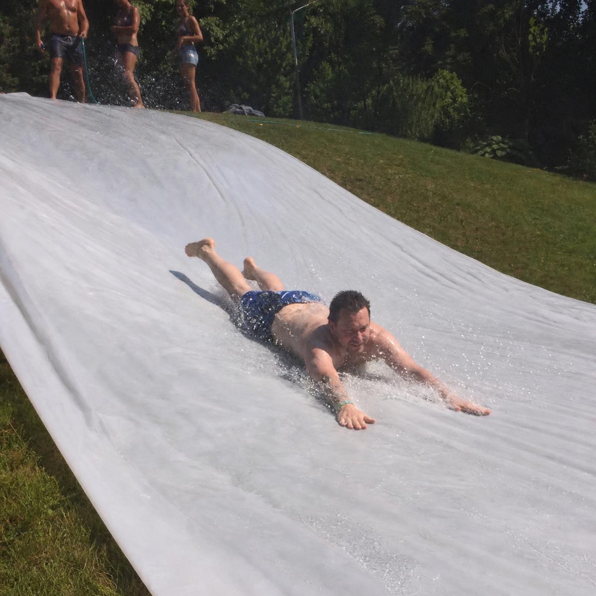An adult man sliding on his belly down a grassy hill on a 100 ft long, 10 ft wide run of soaked plain white plastic slip and slide sheeting