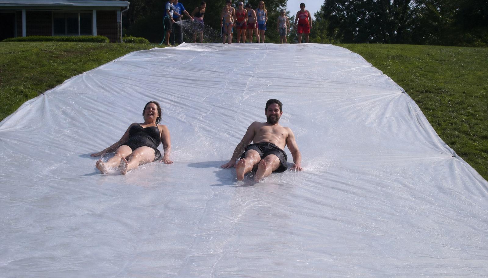 Two smiling adults sliding down a grassy hill side by side on a 100 ft long, 10 ft wide run of soaked plain white plastic slip and slide