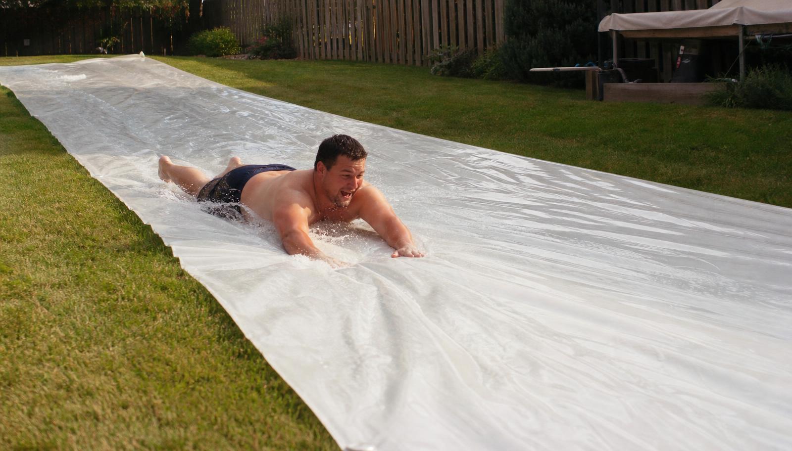 50 Ft Slip and Slide: an adult sliding down a soaked, 10 ft wide run of plain white plastic slip and slide sheeting